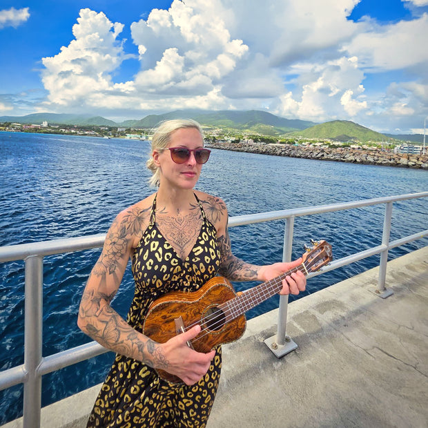 A woman in a dress and wearing sunglasses playing ukulele by the ocean
